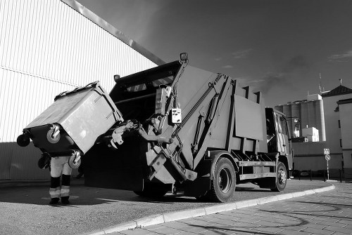 A large industrial rubbish collection truck with a hydraulic arm and compactor mechanism is positioned on a paved outdoor area next to a tall, corrugated metal fence. The truck is predominantly dark with reflective surfaces and has a large, rectangular compaction container at the rear, which is open at the top. In front of the vehicle, a worker wearing striped high-visibility work trousers is seen loading a sizable plastic waste bin, tilted towards the truck’s opening. The bin is light-colored with a textured surface, and the worker is wearing gloves while maneuvering it. The surrounding environment appears to be an industrial or commercial area with a clear sky overhead, and the scene suggests ongoing rubbish removal activity typical of a commercial waste management service in Plaistow. This image illustrates the process of waste collection and material handling relevant to office clearance and rubbish removal services provided by Office Clearance Plaistow, serving the local postcode area.