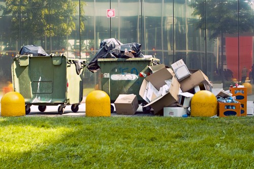 The image displays a row of three large wheelie bins positioned outdoors on a paved area, with two of the bins partially covered by black plastic tarps. To the right of the bins, there is a pile of assorted discarded cardboard boxes, some flattened and others still in their original shape, along with a few plastic bottles and small miscellaneous items. The scene is set against a backdrop of reflective glass windows reflecting trees and a street scene, with a well-maintained grassy verge in the foreground. Bright yellow bollards are situated along the edge of the paved area, providing a safety barrier. The overall environment appears clean and organized, indicative of a location where rubbish or waste is collected and managed, aligning with services offered by Office Clearance Plaistow for rubbish removal in the Plaistow area.