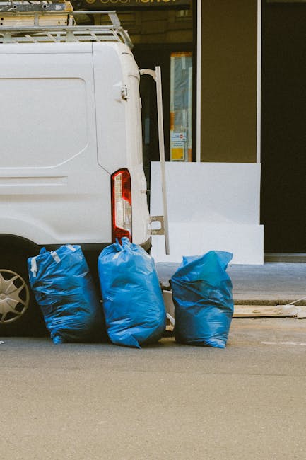 Two waste collection workers are engaged in the process of gathering rubbish in an urban area, standing beside a large, industrial waste disposal vehicle. The worker on the left, wearing a dark green