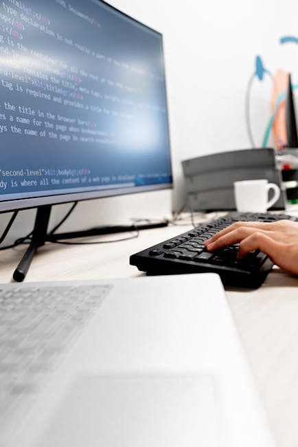 A close-up view of a silver laptop positioned on a light wooden surface, displaying a dark screen with colorful lines of programming code. To the left of the laptop, there is an open spiral-bound note