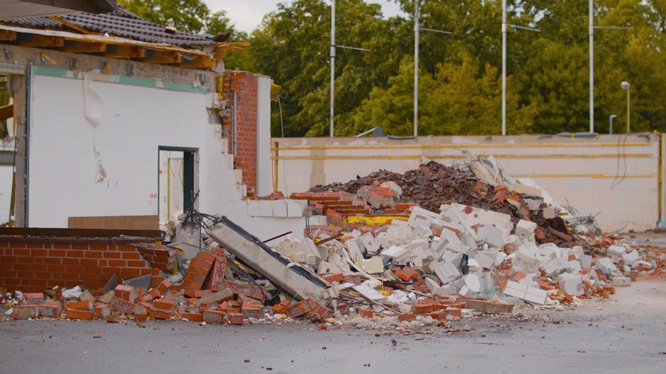 A large pile of construction and demolition debris is situated outdoors on a cleared, paved surface with a light brown, weathered wooden sheet in the foreground. The rubble consists of broken concrete
