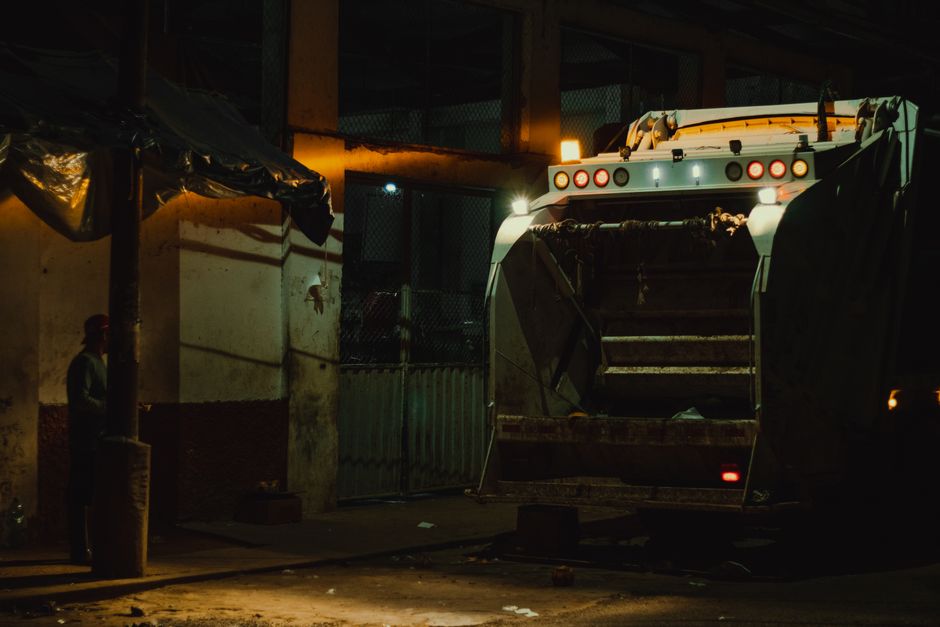 A close-up view of a city refuse collection truck parked on a street, with a visible driver wearing a high-visibility yellow safety vest and glasses operating the vehicle. The truck has a red and whit