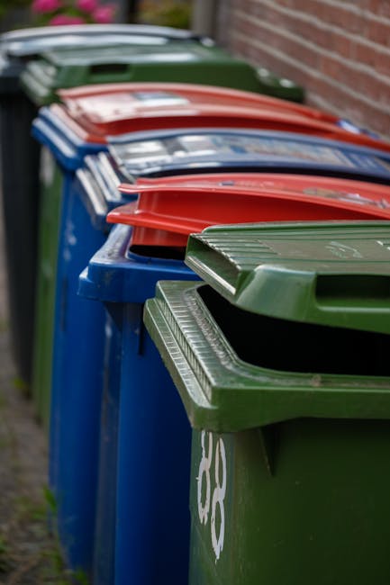 A municipal waste collection worker, dressed in a high-visibility yellow and red uniform, is operating a large red rubbish truck on a roadside, engaged in the process of waste collection. The worker i