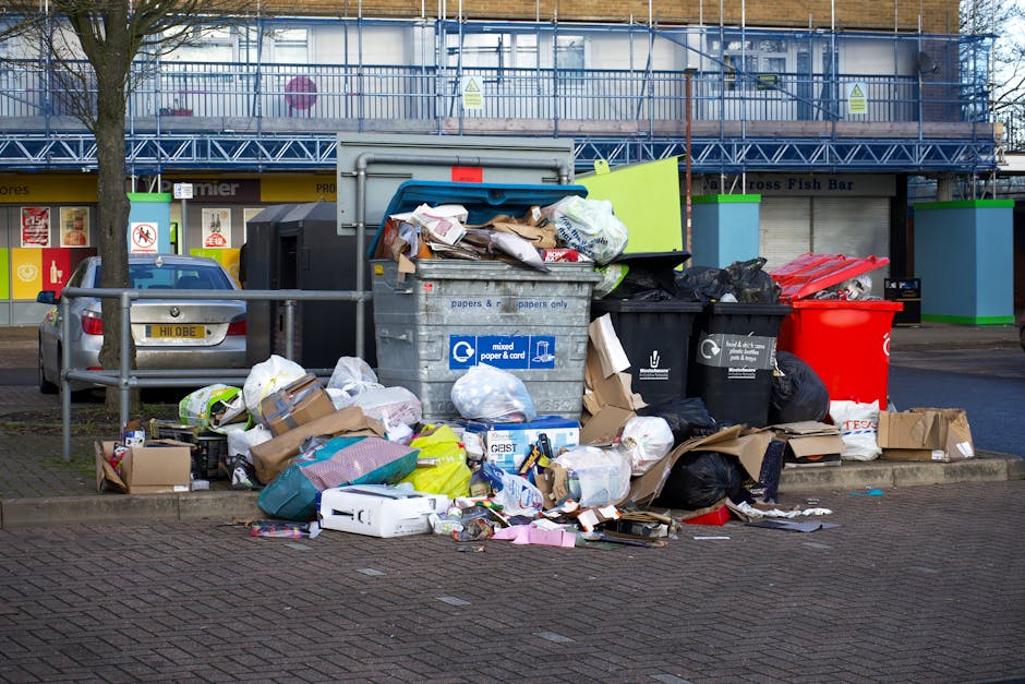 A large pile of transparent plastic garbage bags filled with mixed waste materials, including clothing and packaging, situated on a paved sidewalk in front of a commercial premises at night. Behind th