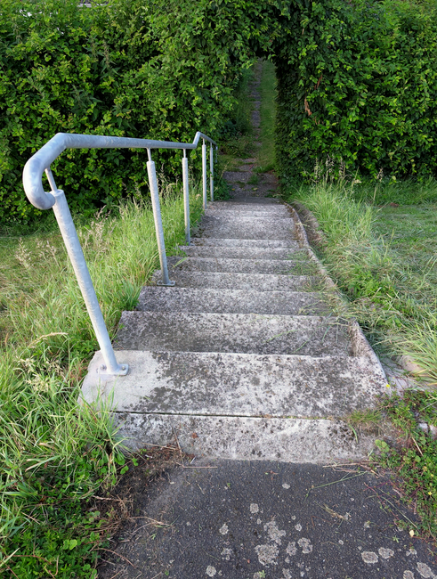 A close-up view of a metal staircase with horizontal, evenly spaced, dark grey metal slats forming the steps. Shadows cast by the railing create a pattern of lines on the steps, which have a slightly 