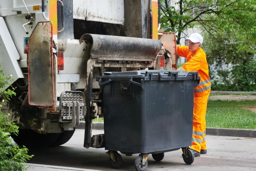 A municipal worker dressed in high-visibility orange overalls and a white cap is standing next to a large black wheeled rubbish bin, positioned on a city street near a kerb. The worker is operating or inspecting the mechanical compactor of a waste collection vehicle, which is partially visible with its rear section and mechanical components. The bin is made of durable plastic, with a textured surface, and is placed in front of the vehicle's loading area. In the background, there are trees and green foliage, suggesting a park or residential area, and a section of pavement can be seen beside the bin. The scene is outdoors during daytime, with natural light illuminating the area, typical for a routine rubbish collection or clearance operation, which is relevant to services offered by Office Clearance Plaistow in the local Plaistow district to manage waste removal efficiently.