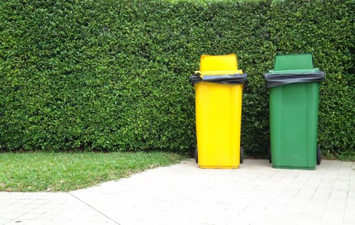 Two large wheelie bins positioned on a paved pathway in front of a dense, well-maintained hedge. The bin on the left is bright yellow with a black lid, while the bin on the right is green with a matching lid. Both bins are closed and appear to be used for domestic rubbish collection, with some black rubbish bags stacked on top of each lid. The pathway is made of light-colored paving stones, and the greenery suggests an outdoor setting typical of residential or commercial premises in Plaistow. The image is clear, capturing the textured surfaces of the plastic bins, the smooth finish of the lids, and the lush, leafy background, illustrating waste disposal equipment ready for collection by a local rubbish removal service such as Office Clearance Plaistow.